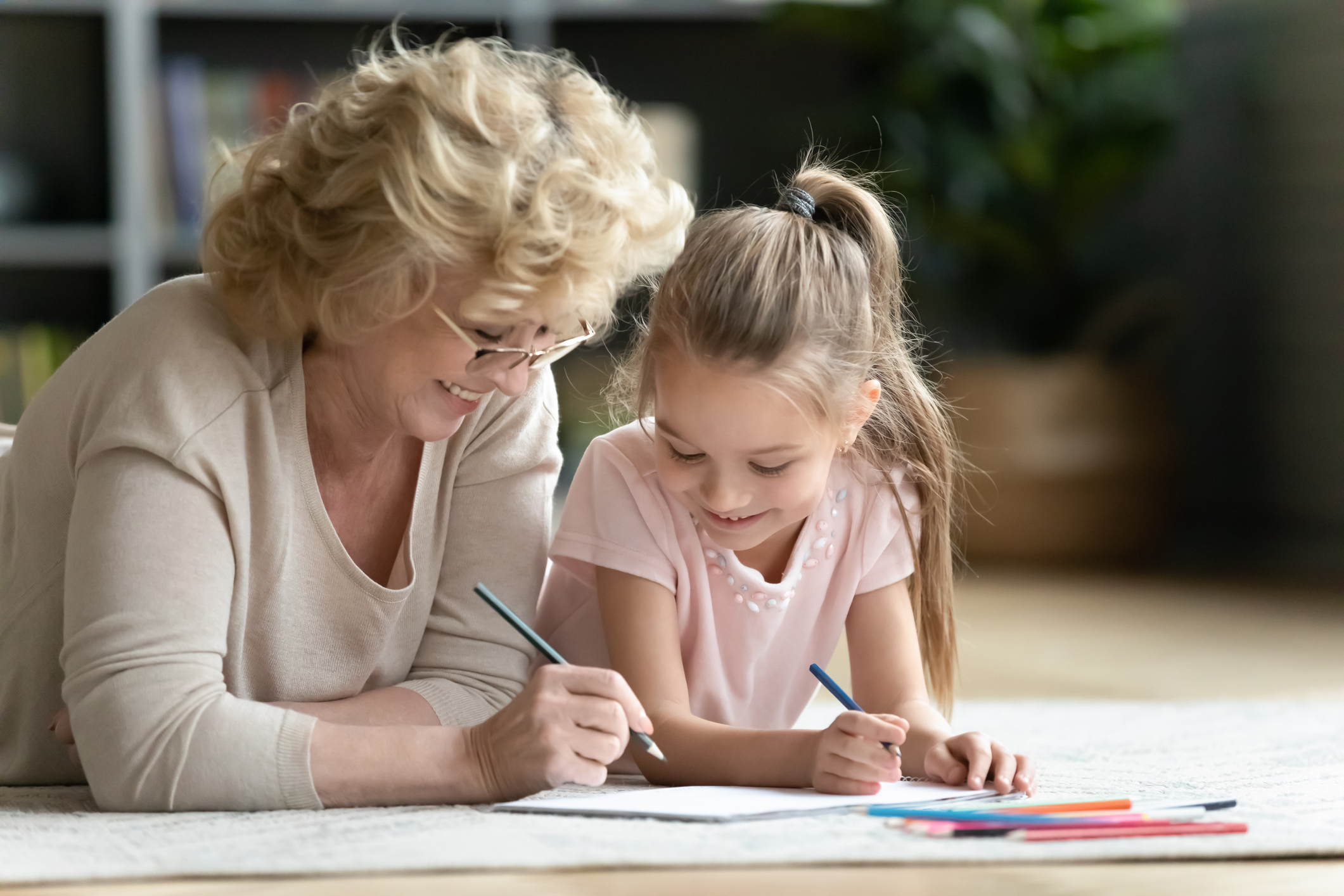 Head shot caring loving older granny or nanny lying on floor with cute small kid girl, drawing pictures in paper album. Affectionate elderly woman teaching painting little preschool granddaughter.
